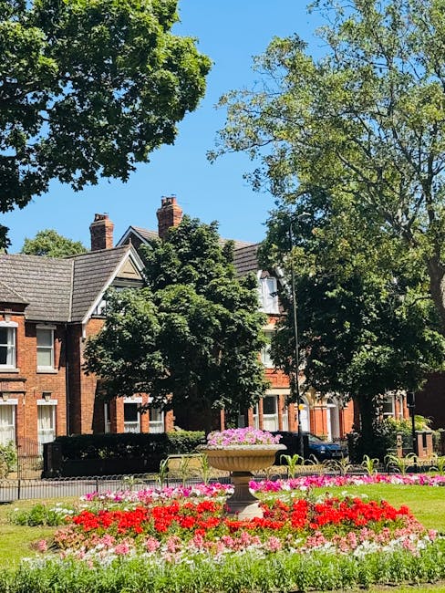 A well-maintained front garden features a vibrant flower bed with red, pink, and white blooms arranged in neat rows, contrasting with the lush green lawn and surrounding trees. A large decorative stone planter, filled with pink flowering plants, sits centrally amid the flower beds. Mature trees with dense foliage provide shade and add height to the garden, their textured bark and broad leaves visible under a bright blue sky. In the background, classic brick houses with sloped tiled roofs, chimneys, and white-framed windows are partially obscured by the trees. The garden area is enclosed by a low black metal fence, and the overall scene suggests recent garden maintenance or landscaping work by Gardeners Uxbridge, with the grass neatly trimmed and the flower beds carefully tended under natural daylight.