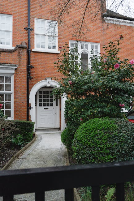 A well-maintained front garden features a vibrant flower bed with red, pink, and white blooms arranged in neat rows, contrasting with the lush green lawn and surrounding trees. A large decorative stone planter, filled with pink flowering plants, sits centrally amid the flower beds. Mature trees with dense foliage provide shade and add height to the garden, their textured bark and broad leaves visible under a bright blue sky. In the background, classic brick houses with sloped tiled roofs, chimneys, and white-framed windows are partially obscured by the trees. The garden area is enclosed by a low black metal fence, and the overall scene suggests recent garden maintenance or landscaping work by Gardeners Uxbridge, with the grass neatly trimmed and the flower beds carefully tended under natural daylight.